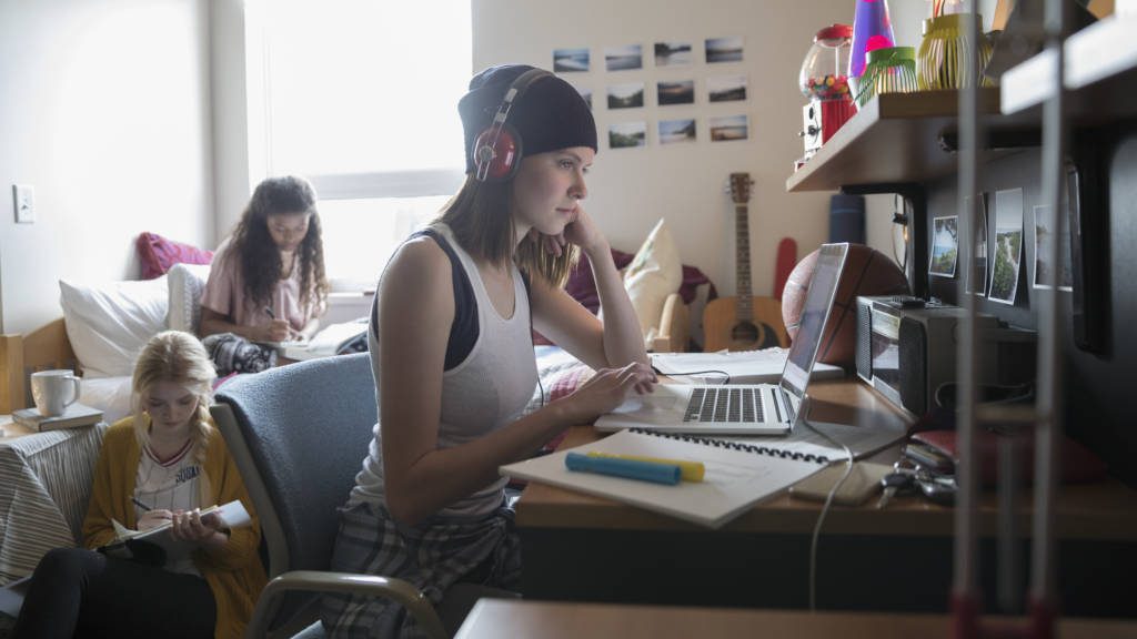 Three college students in a dorm room studying for class, including one working on a laptop and two writing in notebooks