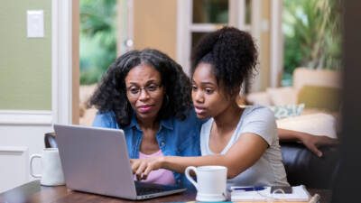 Young woman and her mature mom look at something on a laptop as the young woman studies for a college exam.